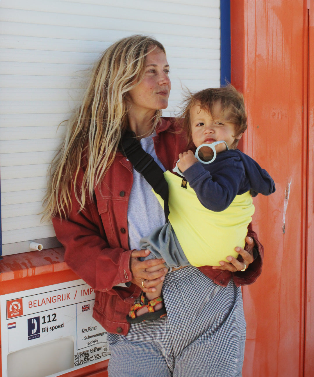 A woman on a beachy scene,  holding her baby with support from the Wildride toddler hip sling carrier, in lemon yellow