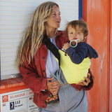 A woman on a beachy scene,  holding her baby with support from the Wildride toddler hip sling carrier, in lemon yellow