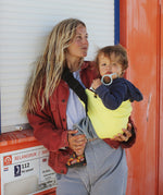 A woman on a beachy scene,  holding her baby with support from the Wildride toddler hip sling carrier, in lemon yellow