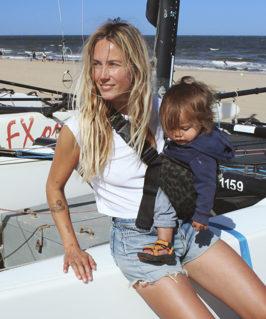 A woman leaning against a yacht, carrying her baby in the stylish Wildride hip sling carrier in grey leopard print