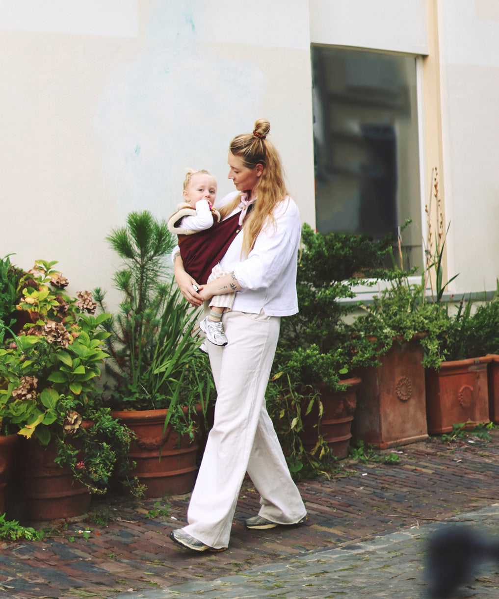 A woman walking down a street, carrying her baby in the Wildride toddler hip sling carrier, in merlot maroon colour