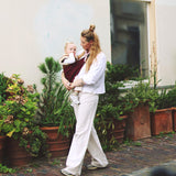 A woman walking down a street, carrying her baby in the Wildride toddler hip sling carrier, in merlot maroon colour