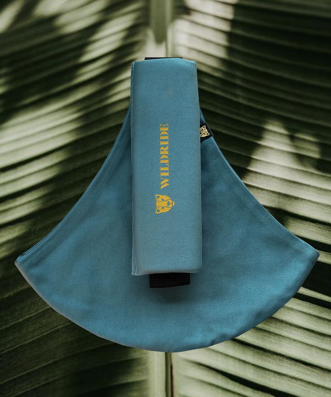 Wildride toddler carrier in ocean. A 100% cotton seat stylish hip carrier for babies with brand name in yellow on the strap that's folded over the carrier's seat with a big green leaf in the background.