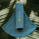 Wildride toddler carrier in ocean. A 100% cotton seat stylish hip carrier for babies with brand name in yellow on the strap that's folded over the carrier's seat with a big green leaf in the background.