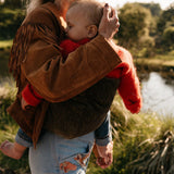 A woman carrying her toddler in the Wildride olive green corduroy toddler carrier, with a nature background featuring a lake.