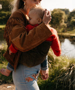 A woman carrying her toddler in the Wildride olive green corduroy toddler carrier, with a nature background featuring a lake.