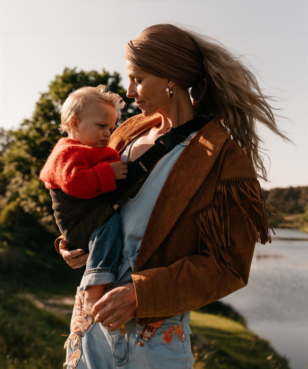 A woman carrying her toddler in the Wildride olive corduroy toddler carrier, with a river in the background.