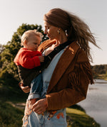 A woman carrying her toddler in the Wildride olive corduroy toddler carrier, with a river in the background.