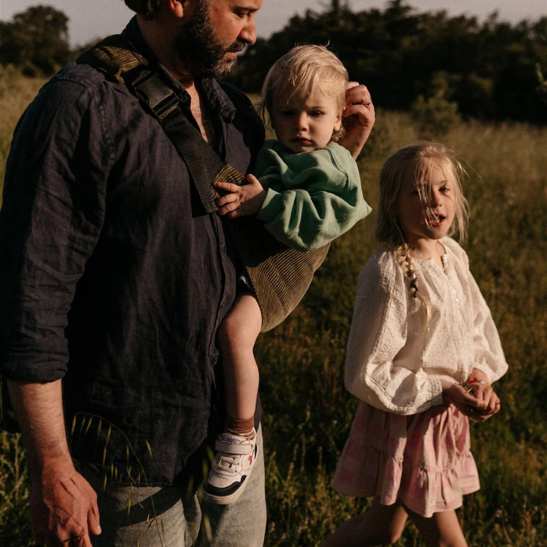 A man carrying his toddler in the olive green corduroy Wildride carrier sling, with his daughter walking next to them.