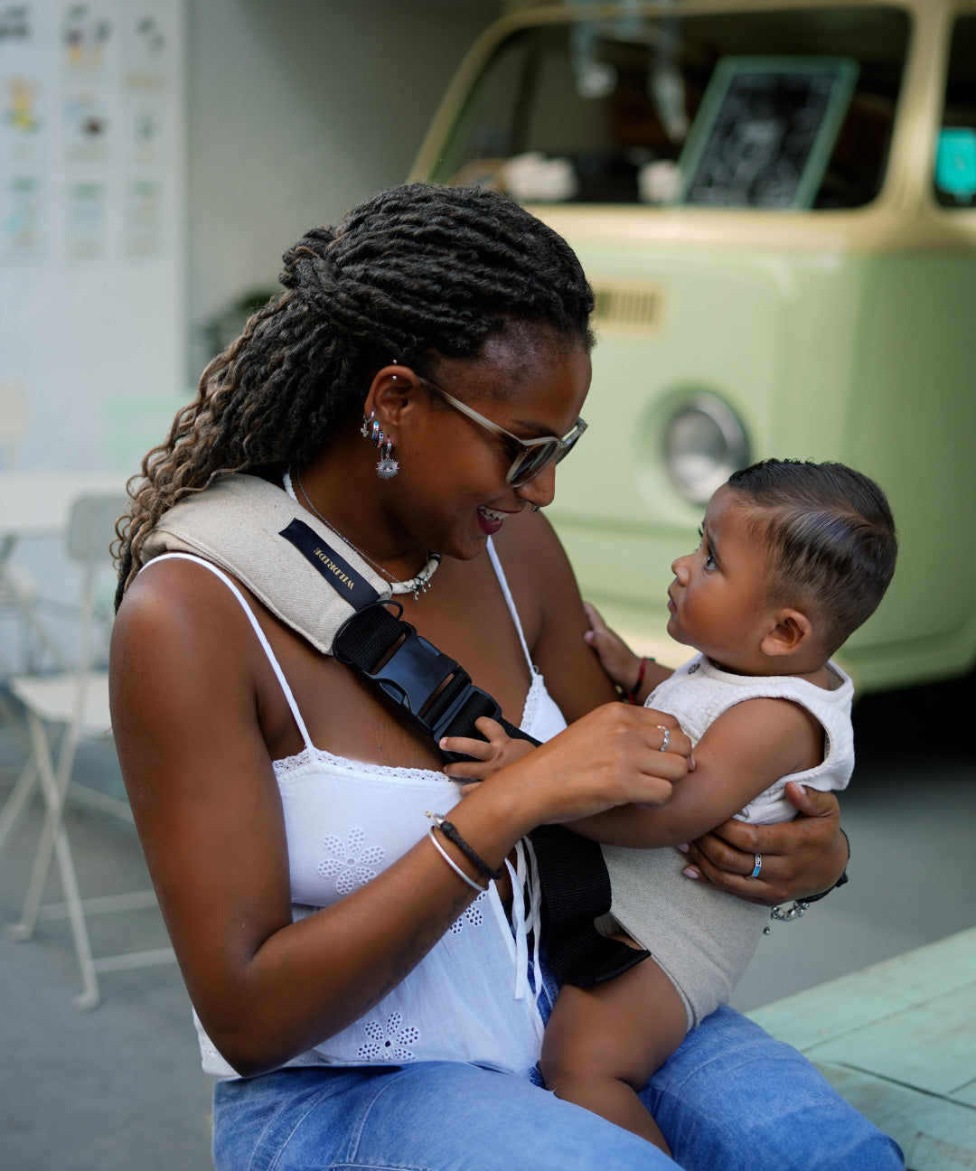 A woman sat down, and supporting her baby with the Wildride ergonomic baby and toddler carrier sling, in front of a vintage camper van.