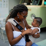 A woman sat down, and supporting her baby with the Wildride ergonomic baby and toddler carrier sling, in front of a vintage camper van.