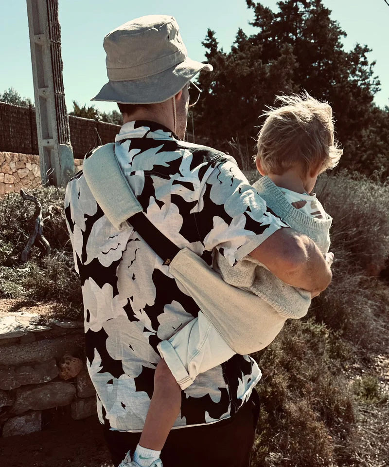 A father carrying a toddler outdoors in the sand linen Wildride toddler carrier showing the detail on the back of the strap