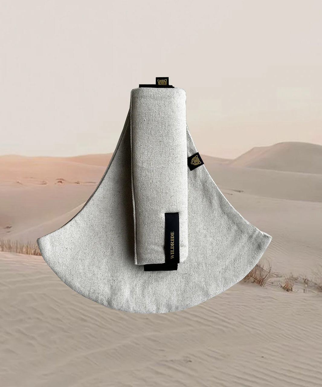 Wildride toddler carrier in sand linen. A stylish hip carrier for babies with a black brand patch on the strap that's folded over the carrier's seat with a desert in the background.