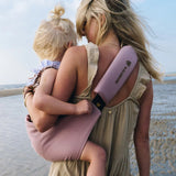A mother carrying a toddler on the beach in the vintage rose Wildride toddler carrier with a child sitting on the seat and showing the detail on the strap and seat