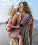 A mother carrying a toddler on the beach in the vintage rose Wildride toddler carrier with a child sitting on the seat and showing the detail on the strap and seat