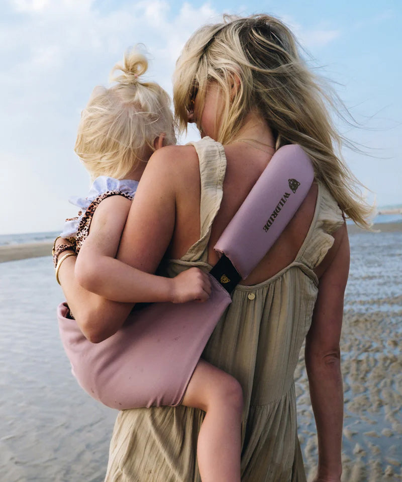 A mother carrying a toddler on the beach in the vintage rose Wildride toddler carrier with a child sitting on the seat and showing the detail on the strap and seat