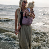 A mother carrying a toddler on the beach in the vintage rose Wildride toddler carrier with a child sitting on the seat and showing the front detail on the strap and seat