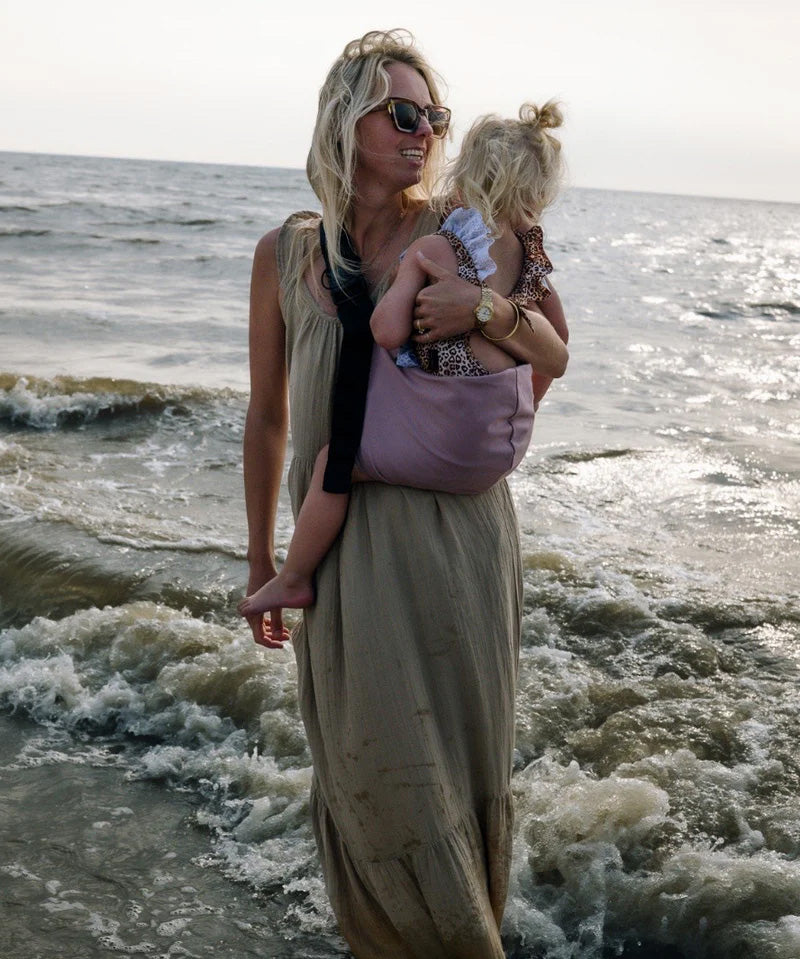 A mother carrying a toddler on the beach in the vintage rose Wildride toddler carrier with a child sitting on the seat and showing the front detail on the strap and seat