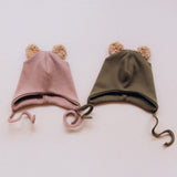 Wooly Organic hat with teddy ears in pale blush / savannah next to the tea leaf green coloured hat both available at Babipur. A GOTS organic hat in a soft ribbed style. Features ties and cute fluffy brown teddy bear ears
