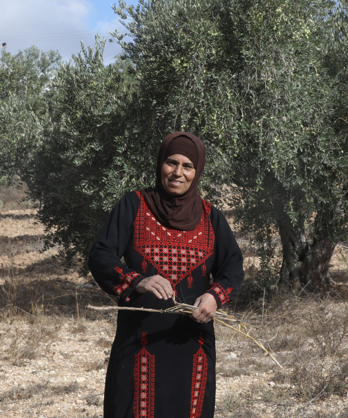 Doha Asous is a family farmer from the ancient Palestinian village of Burin, near Nablus. She is pictures wearing patterned clothing next to an olive tree.