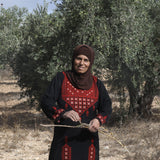 Doha Asous is a family farmer from the ancient Palestinian village of Burin, near Nablus. She is pictures wearing patterned clothing next to an olive tree.