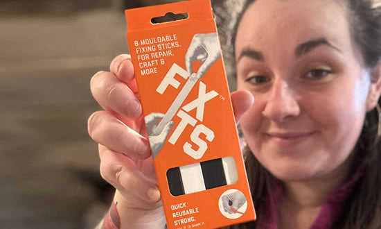 A Caucasian woman with brown hair holds up a small rectangular orange cardboard packet in front of her face. The packet has the word "FIXITS" written in white on it