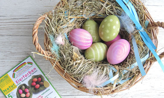  different pastel colors is placed on a pale wooden background. The eggs range from light pink, blue, green, purple, and yellow. To the right of the basket, there is a packet of egg dye, suggesting the process used to achieve the colored eggs