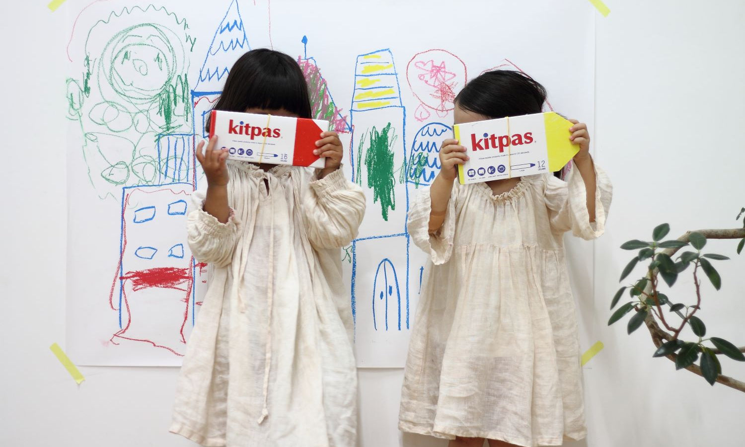 Two children in white dresses holding up a pack of Kitpas Crayons standing in front of a whiteboard with their art work