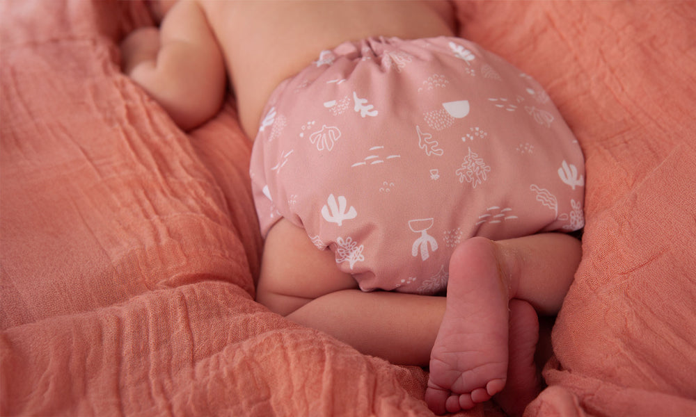 Newborn baby wearing a reusable TotsBots cloth nappy on an orange muslin blanket to represent 'Guide to Newborn Reusable Nappies' blog by Babipur.