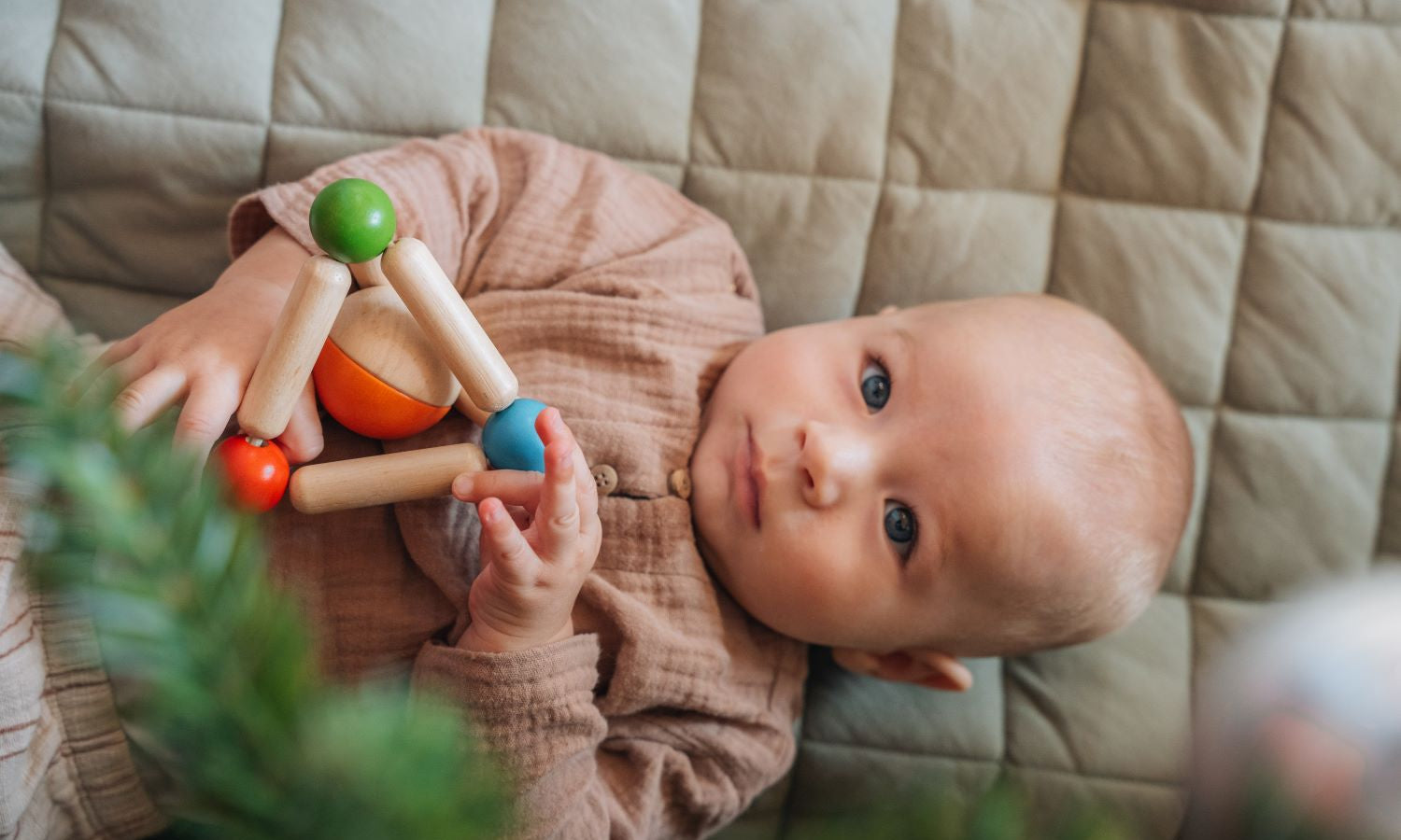 A baby on a blanket looking up through the Christmas Tree holding a wooden toy by PlanToys