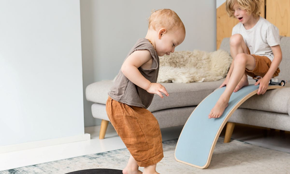 Toddler playing on a Wobbel board starter as a bridge with a sibling in the background with standard wobbel as a slide off the sofa