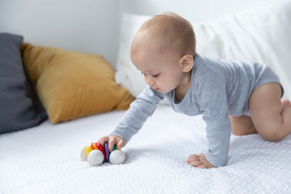 Plan Toys Rainbow Car Rattle Toy is a solid rubber wood sensory toy for babies painted in bright rainbow colours. Baby pushing it along.