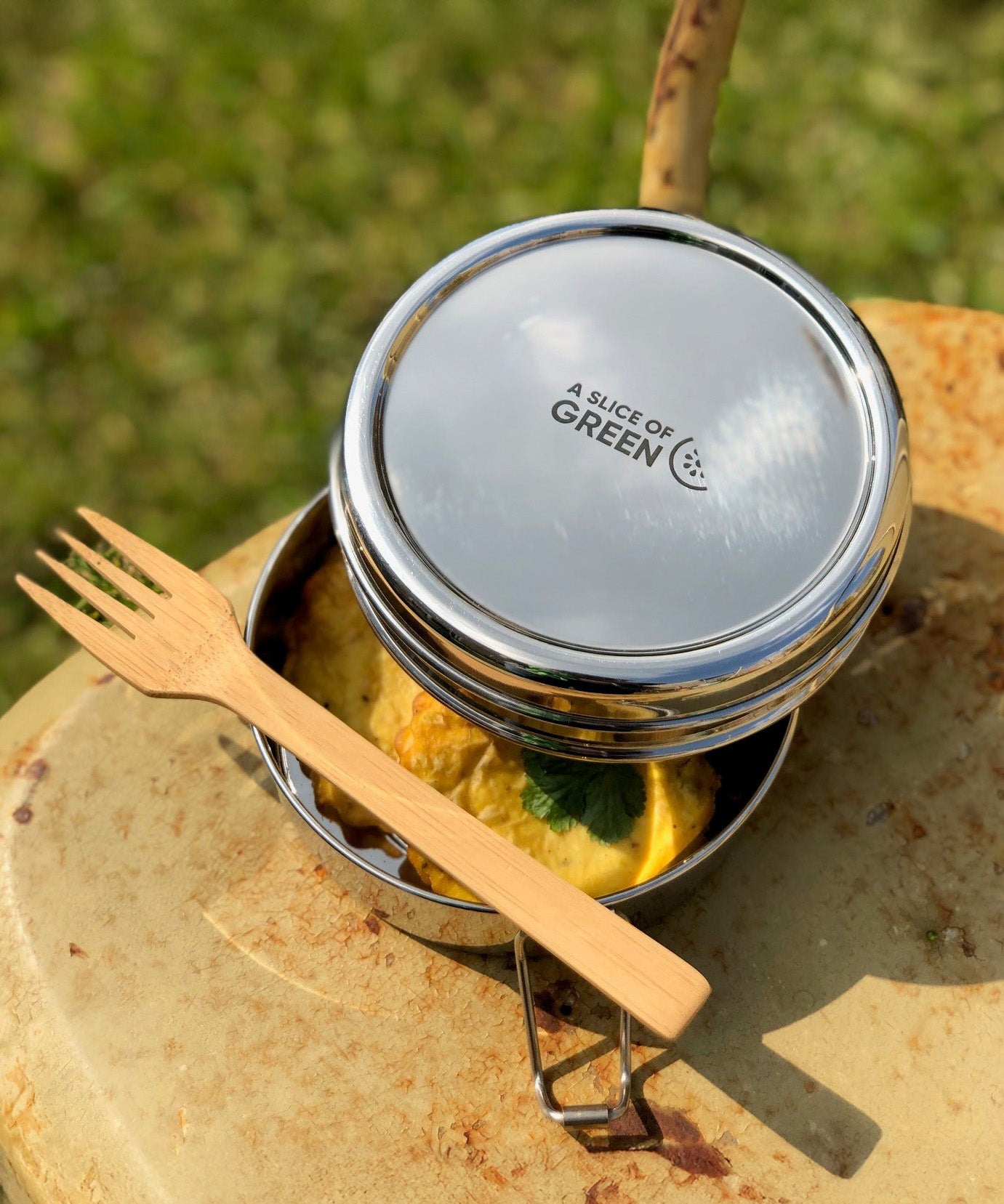 A close up of food items inside one of the tins from the stainless steel A slice of green chapra two tier round lunchbox with a bamboo fork laying on top