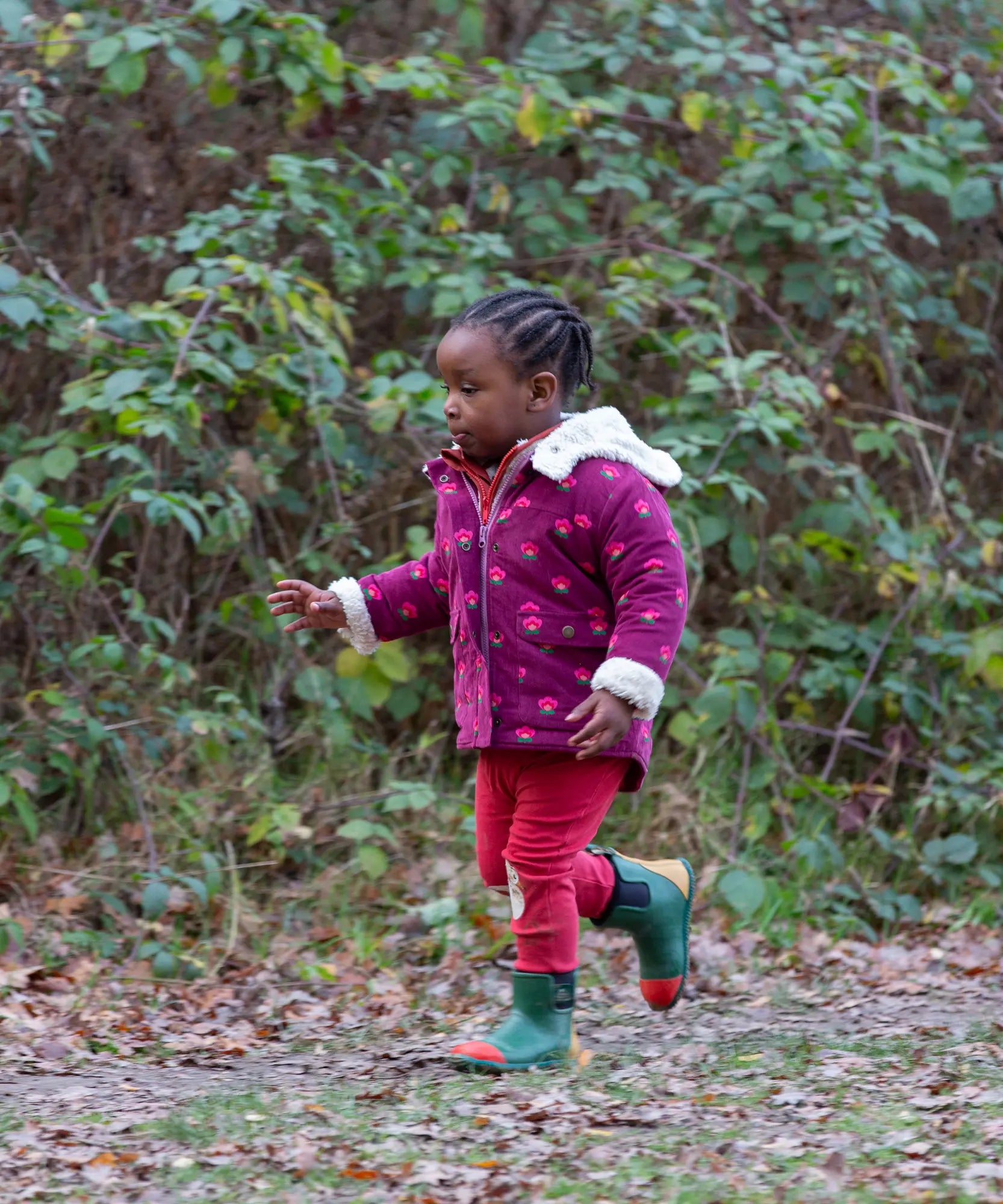 Child wearing a Little Green Radicals pink flowers purple organic cotton sherpa toasty coat with red trousers and green wellington boots.