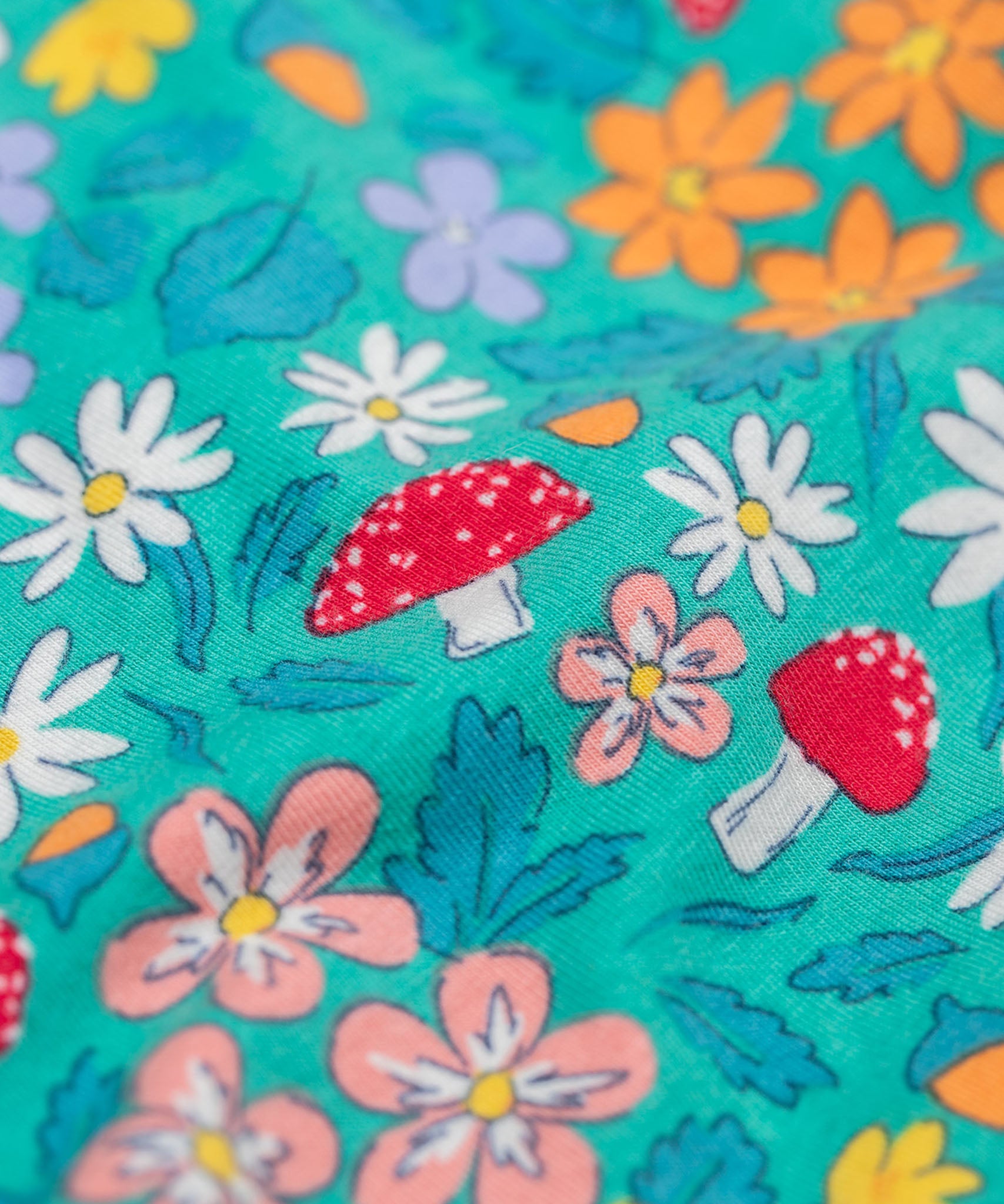 A super close up of the print on the Frugi Sofia skater dress with a wild woods print design on a green background. The print features flowers, toadstools and leaves.