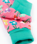A close up of the turquise cloured turn up leg cuffs on the Frugi fun flowers print parsnip pants. The fun flowers print features multi-coloured flowers and bees on a pink background with turquoise coloured waistband and leg cuffs.