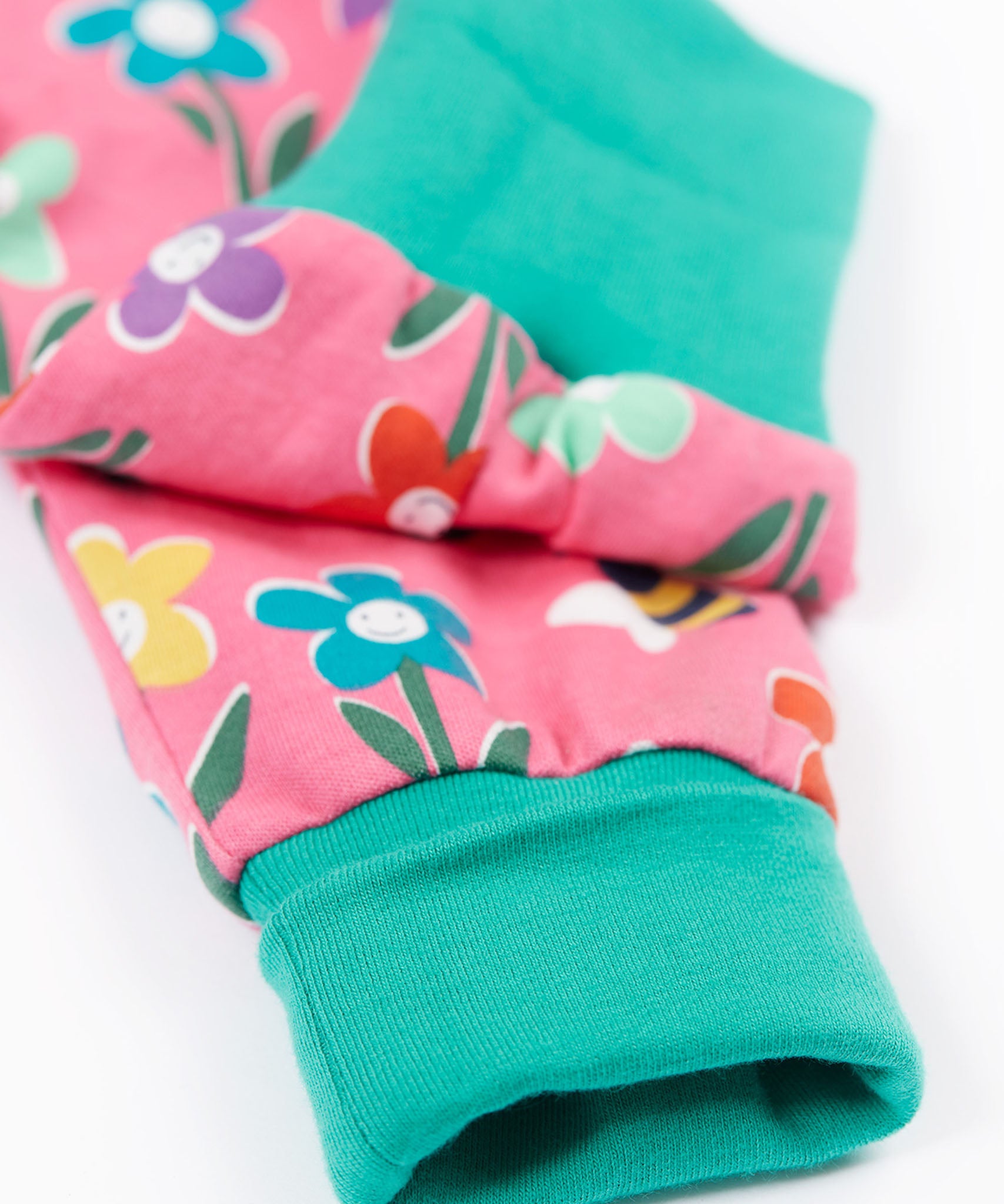 A close up of the turquise cloured turn up leg cuffs on the Frugi fun flowers print parsnip pants. The fun flowers print features multi-coloured flowers and bees on a pink background with turquoise coloured waistband and leg cuffs.