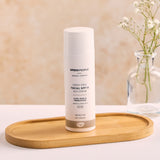 The Green People, Organic, Scent Free Facial SPF15 Sun Cream - 50ml, is stood on a wooden tray on a white table and peach background. There is also a small glass vase with a small plant inside