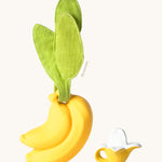 The Ana Banana Pacifier Holder on a cream background. The image shows an Oli & Carol banana teething toy next to the holder. The banana is smooth and has a green muslin leaf attachment on the top to tie the holder to a pram handle, high chair etc.