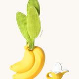 The Ana Banana Pacifier Holder on a cream background. The image shows an Oli & Carol banana teething toy next to the holder. The banana is smooth and has a green muslin leaf attachment on the top to tie the holder to a pram handle, high chair etc.