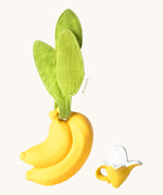 The Ana Banana Pacifier Holder on a cream background. The image shows an Oli & Carol banana teething toy next to the holder. The banana is smooth and has a green muslin leaf attachment on the top to tie the holder to a pram handle, high chair etc.