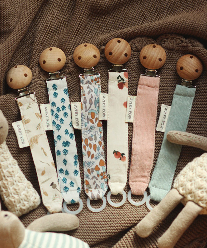 6 Avery Row Pacifier Holders in different colours and prints, on a brown baby blankets and cuddly toys surrounding