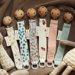 6 Avery Row Pacifier Holders in different colours and prints, on a brown baby blankets and cuddly toys surrounding