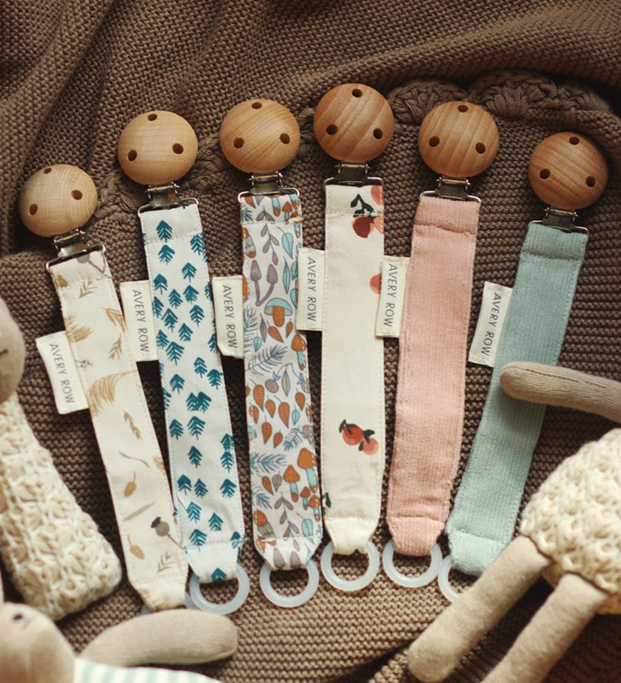 6 Avery Row Pacifier Holders in different colours and prints, on a brown baby blankets and cuddly toys surrounding