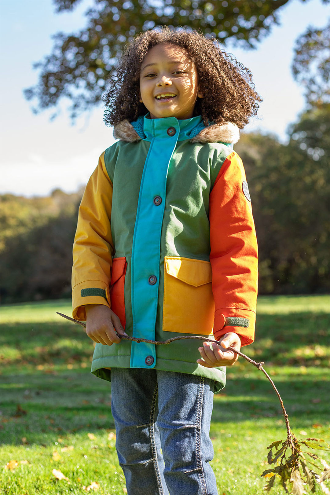 Child wearing a colorful Frugi jacket holding a branch in a park. Representing Mid Season Clothing Sale at Babipur