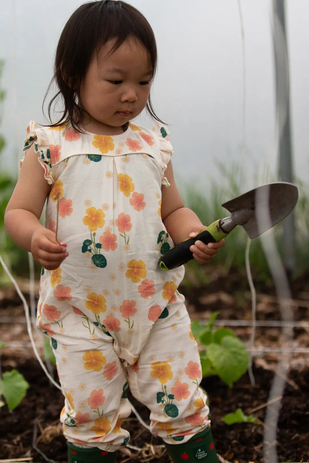 babipur Summer Sale. Little Green radicals organic kids clothing image - A toddler stands in a garden wearing a cream floral jumpsuit with yellow, pink, and green flowers, holding a small hand trowel.