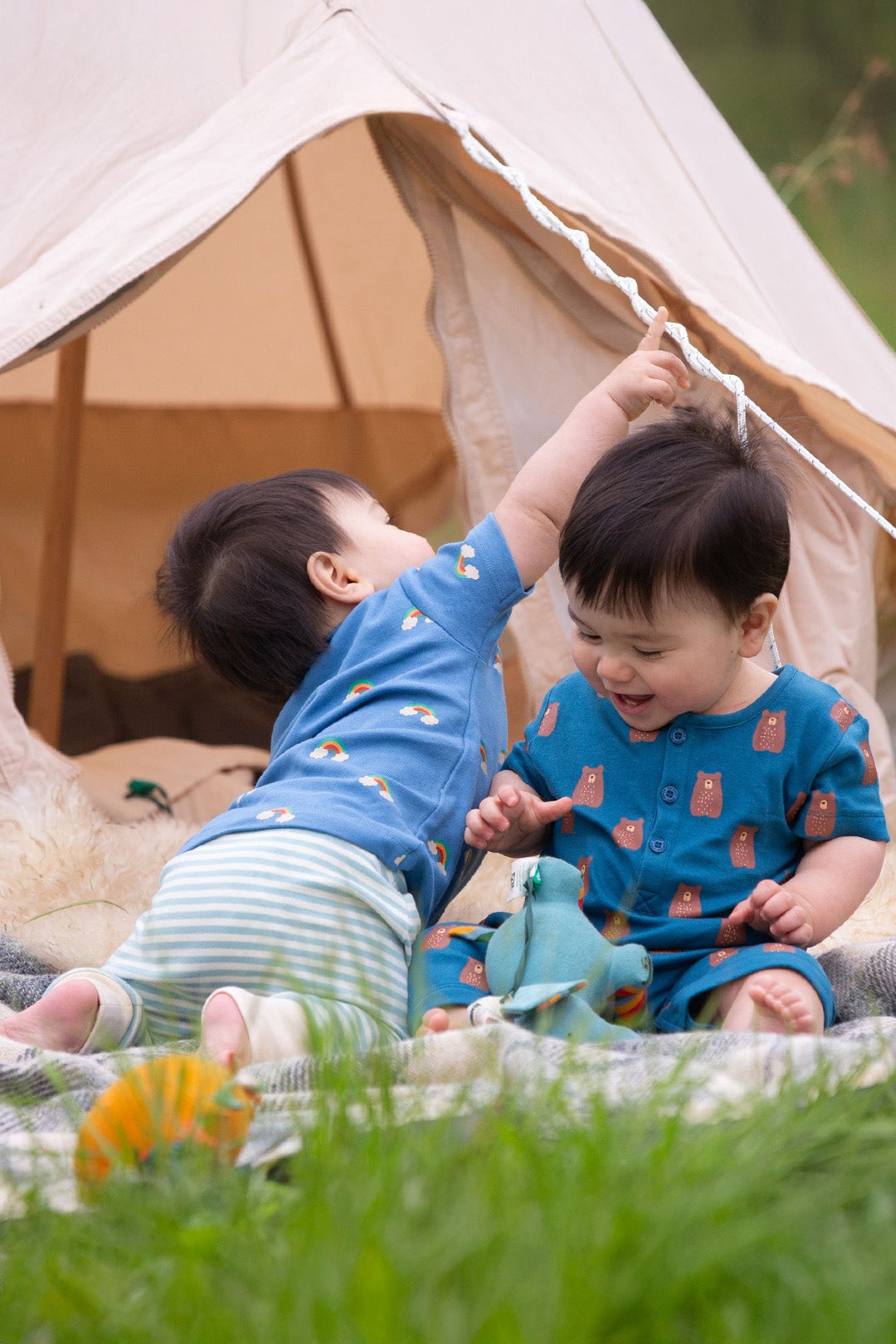 Two toddlers playing wearing Little Green Radicals Organic Clothing with rainbows, stripes and bears.  Available from babipur. Backdrop is outdorrs green and a canvas tent. 