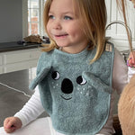 A child sat in a kitchen, wearing the Roommate Organic Cotton Baby Bib - Koala