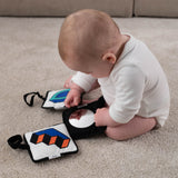 A baby sat down on a soft beige carpet, looking at themselves in the Etta Loves x Camille Walala Sensory Hanging Squares black square with a mirror in the centre