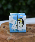 A 8-piece Bajo 2 in 1 cube wooden puzzle featuring penguin illustrations on a blue and white icy theme, displayed on a rustic wooden slice with a blurred green background.